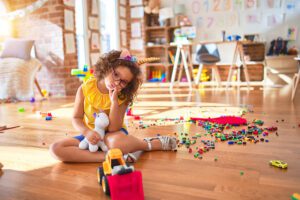 Beautiful toddler wearing glasses and unicorn diadem sitting on the floor at kindergarten thinking looking tired and bored with depression problems with crossed arms.